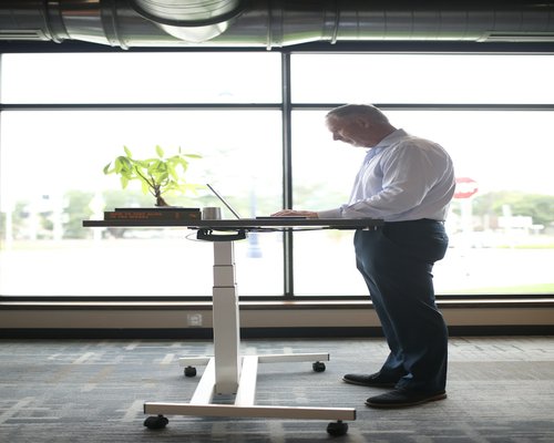 A man standing at a modern standing desk, adjusting his ergonomic chair and screen height